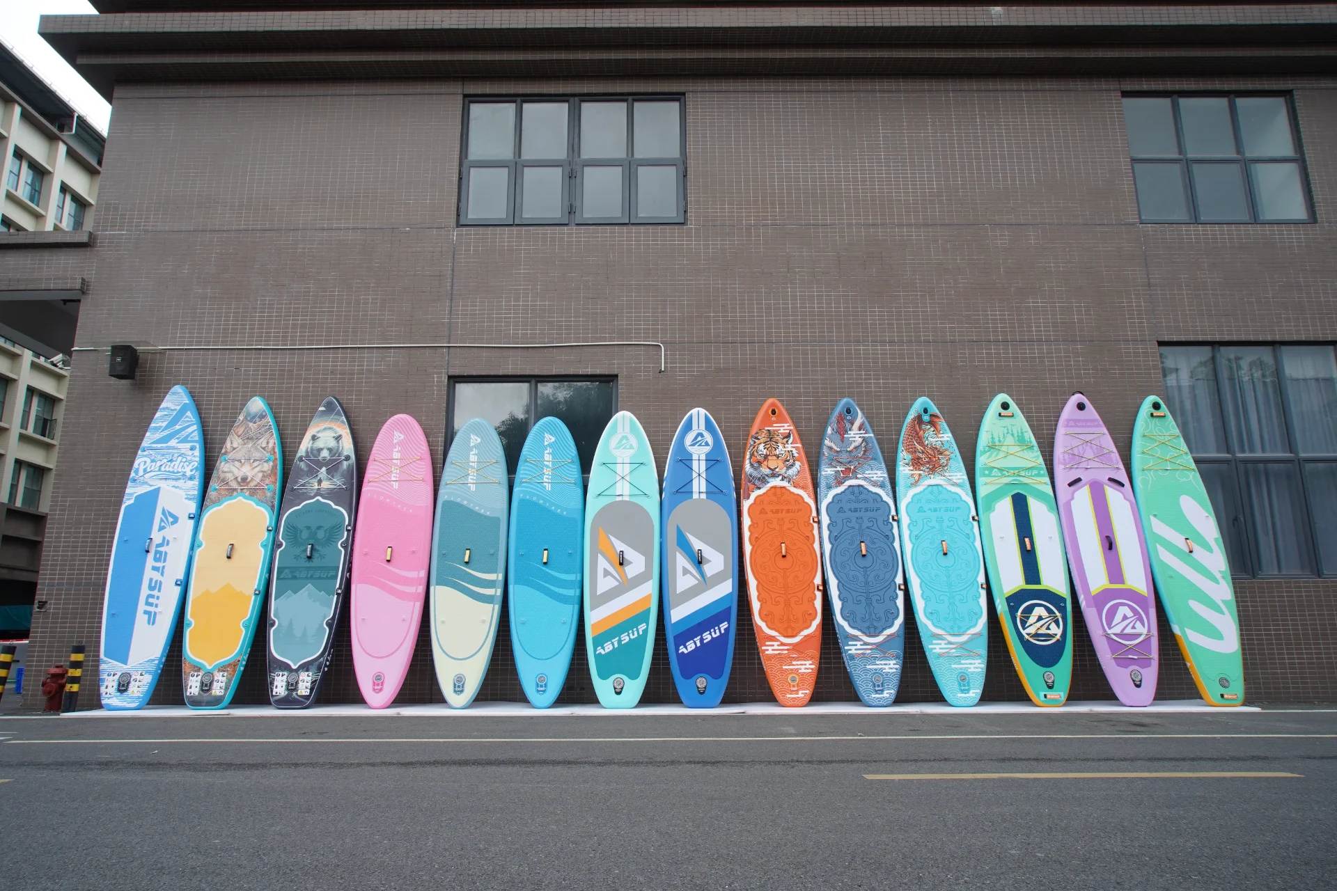 A row of colorful paddleboards with various designs leaning against a building wall on overcast pavement.