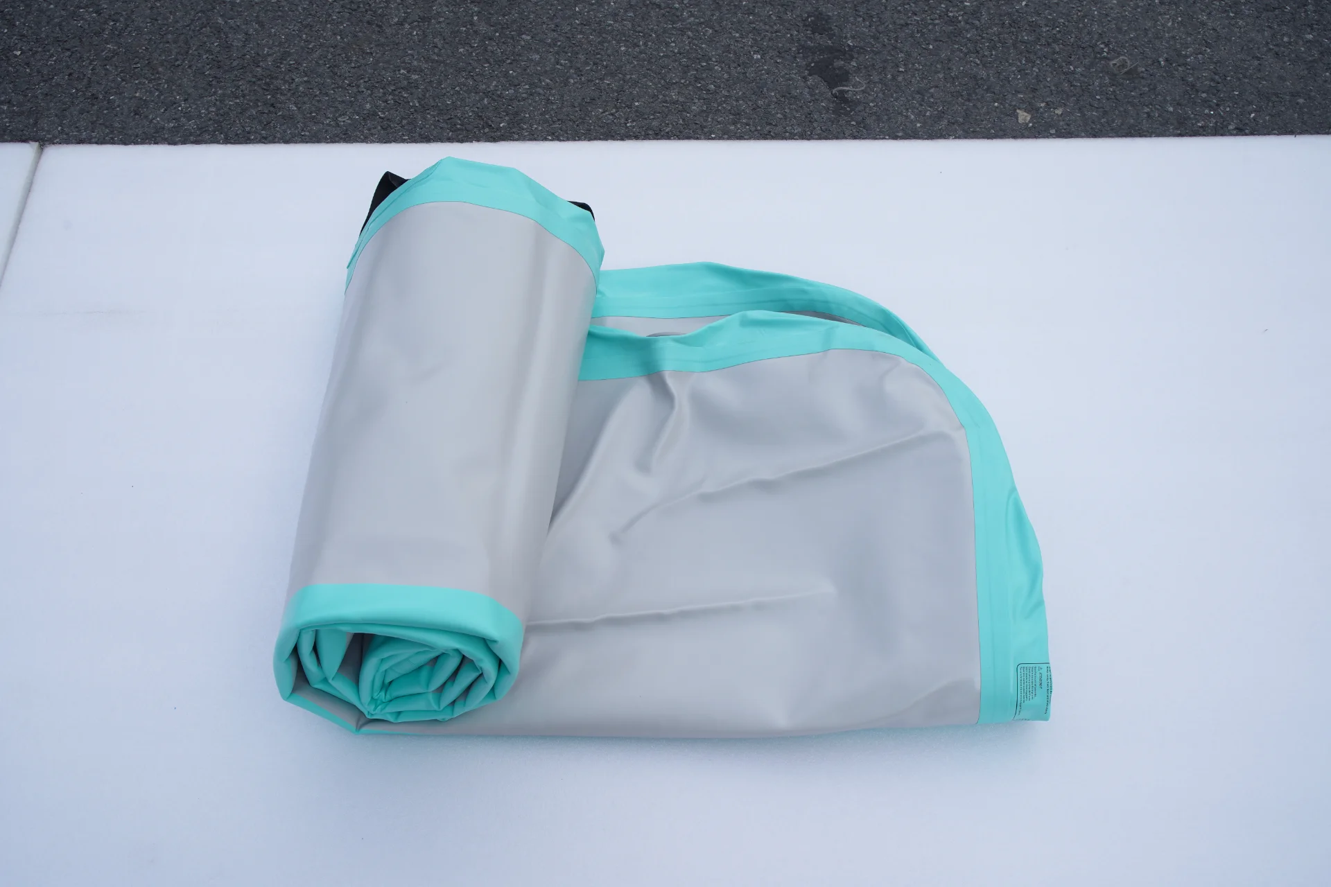 A folded gray and aqua outdoor mat lying on a white surface, with dark pavement visible in the background.
