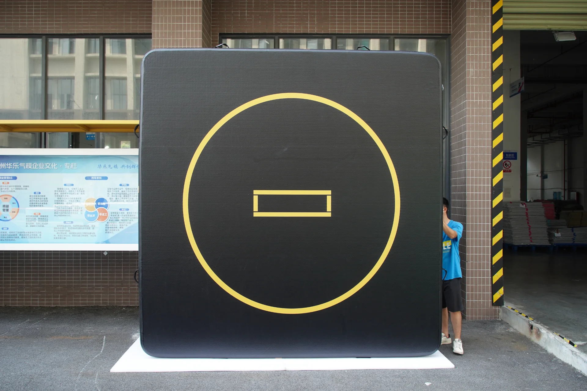 A giant black panel with a yellow circle and horizontal symbol, partially hiding a person, placed outdoors near a building.