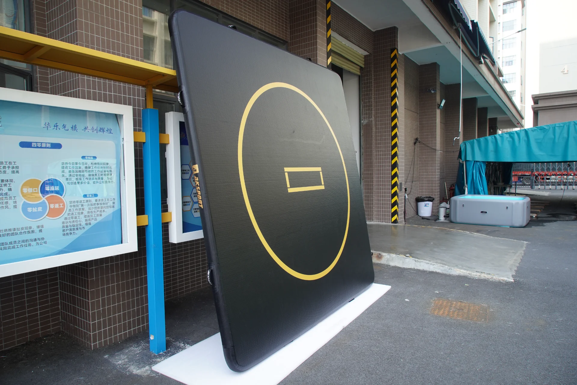 Large black inflatable mat with a yellow circular design leaning against a building in an outdoor setting, near a blue canopy and educational signage.