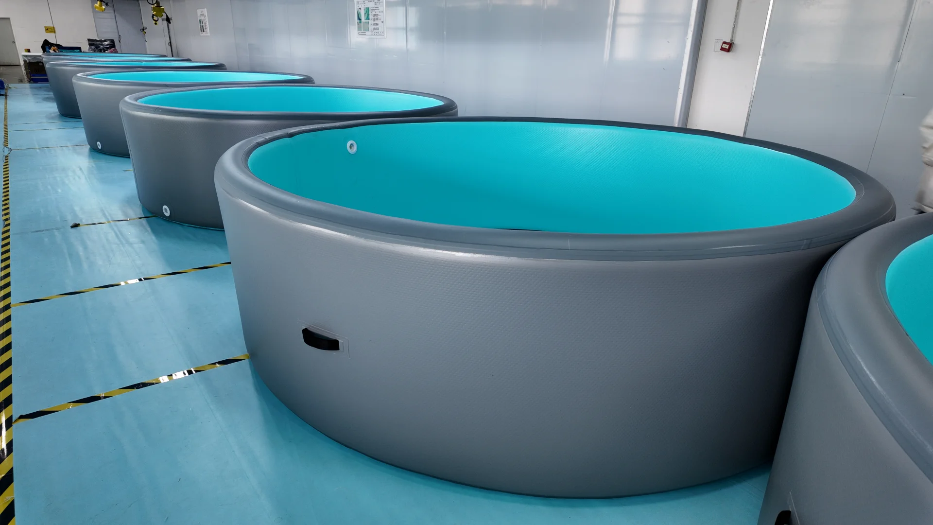 Rows of large gray and blue circular tanks in a clean industrial facility, placed on a blue and yellow-marked floor.