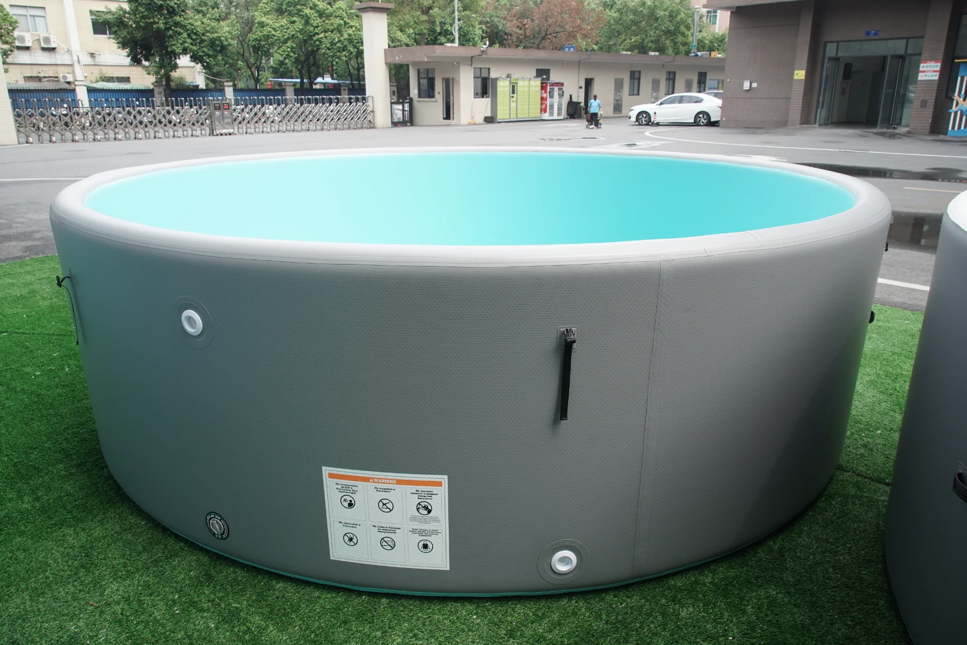 A gray round portable inflatable pool filled with water, placed on a grass surface in an outdoor space with buildings and parked vehicles in the background.