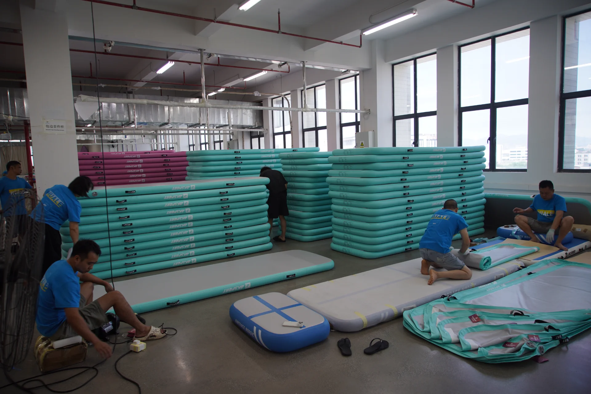 Factory workers assembling and inspecting teal and pink inflatable mats, with stacks of completed mats in the background.
