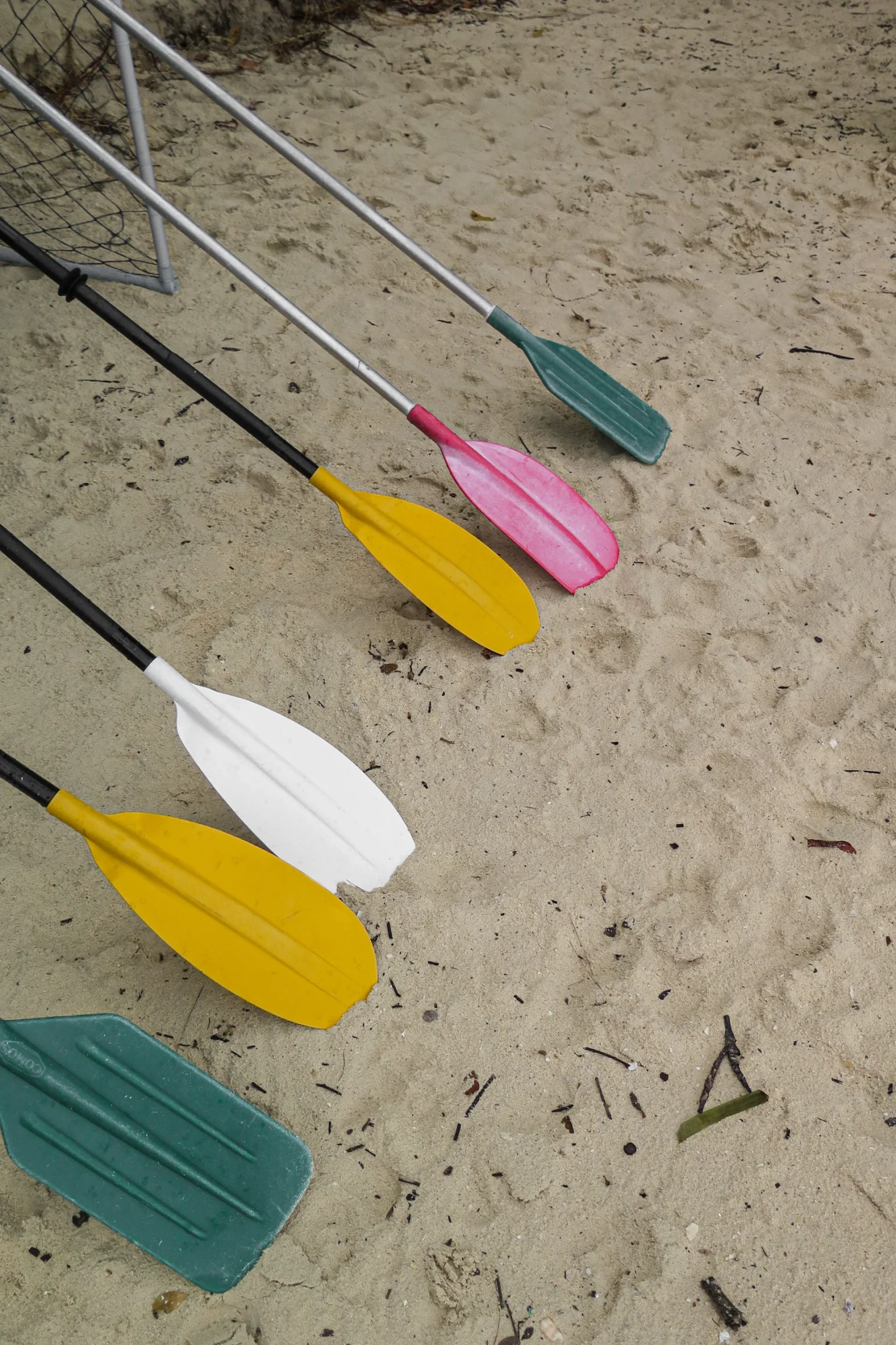 A set of colorful paddles, including yellow, white, pink, and green, lying on a sandy beach surface near a netted frame.