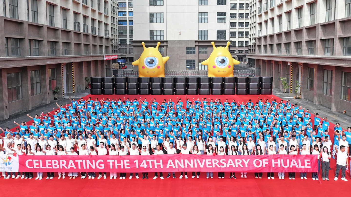 A large group celebrating the 14th anniversary of Huale with participants holding a red banner surrounded by buildings and decorations.