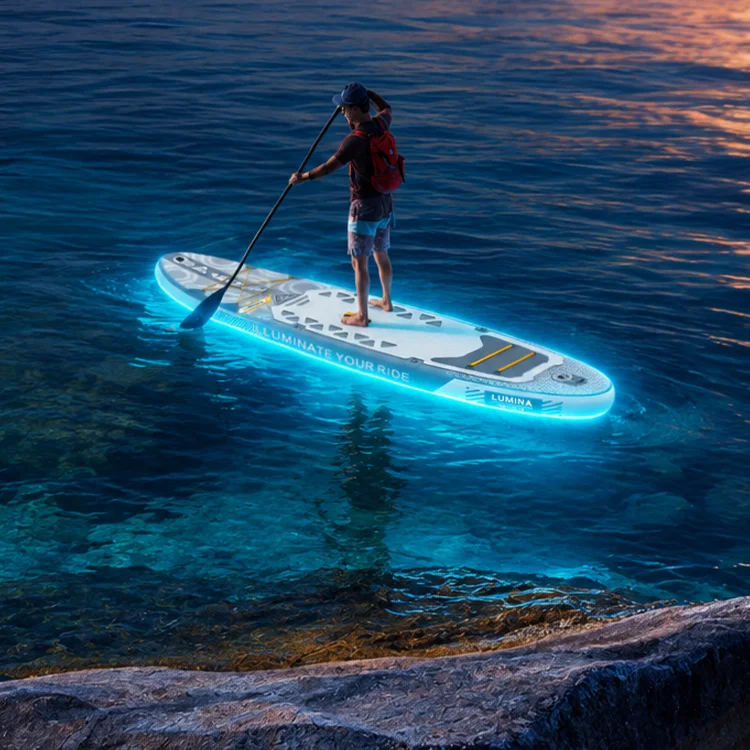 A person standing on an illuminated paddleboard with a paddle, wearing a red backpack, on calm water during twilight.