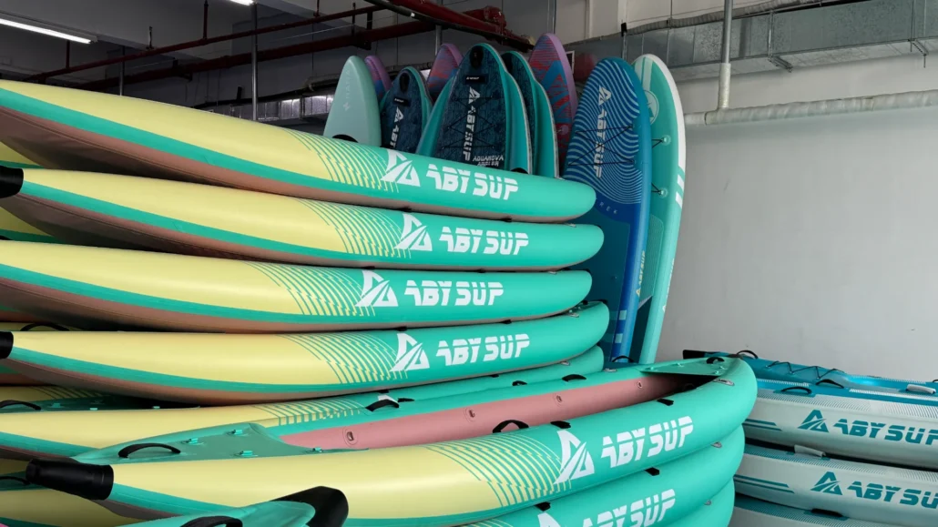 Multiple stacked paddleboards with colorful designs and ABY SUP branding, stored indoors against a wall.