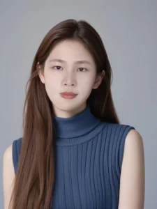 Young woman with long dark hair wearing a blue ribbed turtleneck top, posing against a neutral gray background.