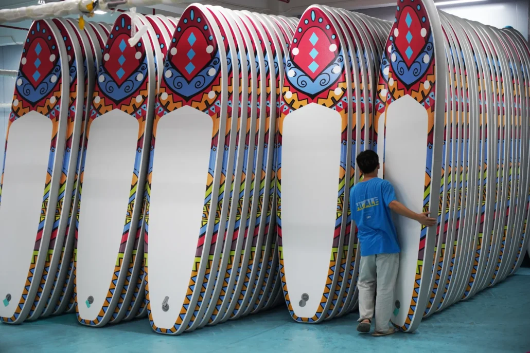 A worker in a blue shirt arranging a stack of colorful paddleboards with vibrant geometric patterns in a factory setting.